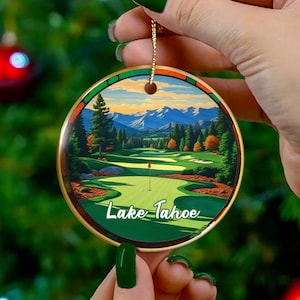 May include: Round ceramic ornament featuring a golf course scene with the text "Lake Tahoe." The ornament has a gold-colored trim and a gold string for hanging. The background is a Christmas tree with red ornaments.