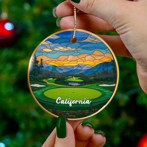 May include: A round Christmas ornament with a golf course scene. The ornament has a gold border and a string for hanging. The image features a green golf course with the word "California" in white. The background shows a sunset with blue, orange, and yellow hues.