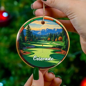 May include: A round Christmas ornament featuring a golf course scene with the word "Colorado" in white. The ornament has a gold-colored trim and a gold string for hanging. The background shows a green Christmas tree with red ornaments.