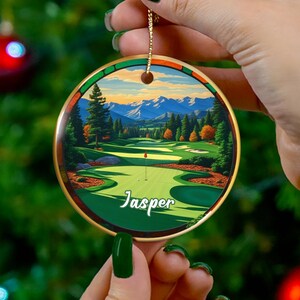 May include: A round ceramic ornament with a golf course scene, featuring green fairways, trees, and mountains under a blue sky. The word "Jasper" is written on the ornament. The ornament has a gold-colored edge and a gold string for hanging.