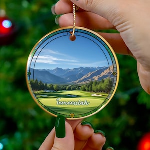 May include: A round, gold-rimmed Christmas ornament featuring a scenic golf course landscape with mountains and blue skies. The word "Temeculah" is printed on the ornament. The ornament is held by a person in front of a Christmas tree.