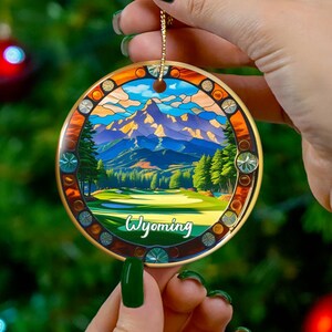 May include: A round Christmas ornament featuring a landscape of mountains, trees, and a lake, with the word "Wyoming" in a script font. The ornament has a gold-colored border and a gold string for hanging. The background is a Christmas tree with red ornaments.