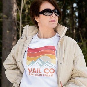 May include: A woman wearing a white sweatshirt with a retro-style graphic that says "Vail Co. Elevation 8022 ft" and a beige quilted jacket. She is standing in a snowy landscape with trees in the background.