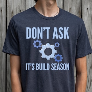 Op de afbeelding: Een donkerblauw t-shirt met de tekst "Don't Ask It's Build Season" en een afbeelding van blauwe tandwielen.