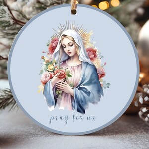 May include: A blue and white Christmas ornament featuring a watercolor painting of the Virgin Mary holding a bouquet of pink and yellow roses. The ornament has the text "pray for us" at the bottom.
