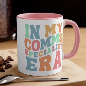May include: A white ceramic mug with a pink rim and handle. The mug has a colorful graphic that says "In My Comms Specialist Era".