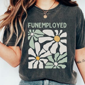 May include: A dark gray t-shirt with a graphic print of stylized white daisies with green leaves and the text "FUNEMPLOYED" in white.