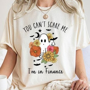 May include: A cream-coloured t-shirt with a Halloween graphic. The design includes a ghost, flowers, bats, and the text "You can't scare me, I'm in Finance." The shirt is a casual, short-sleeved style.