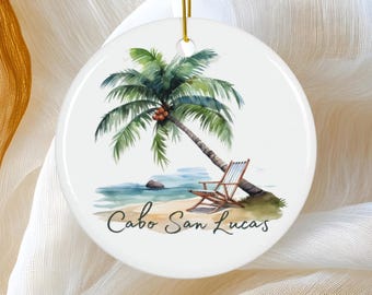 May include: A round, white ceramic ornament featuring a watercolor illustration of a beach scene. A palm tree, a beach chair, and the text "Cabo San Lucas" are depicted. A gold string is attached for hanging.