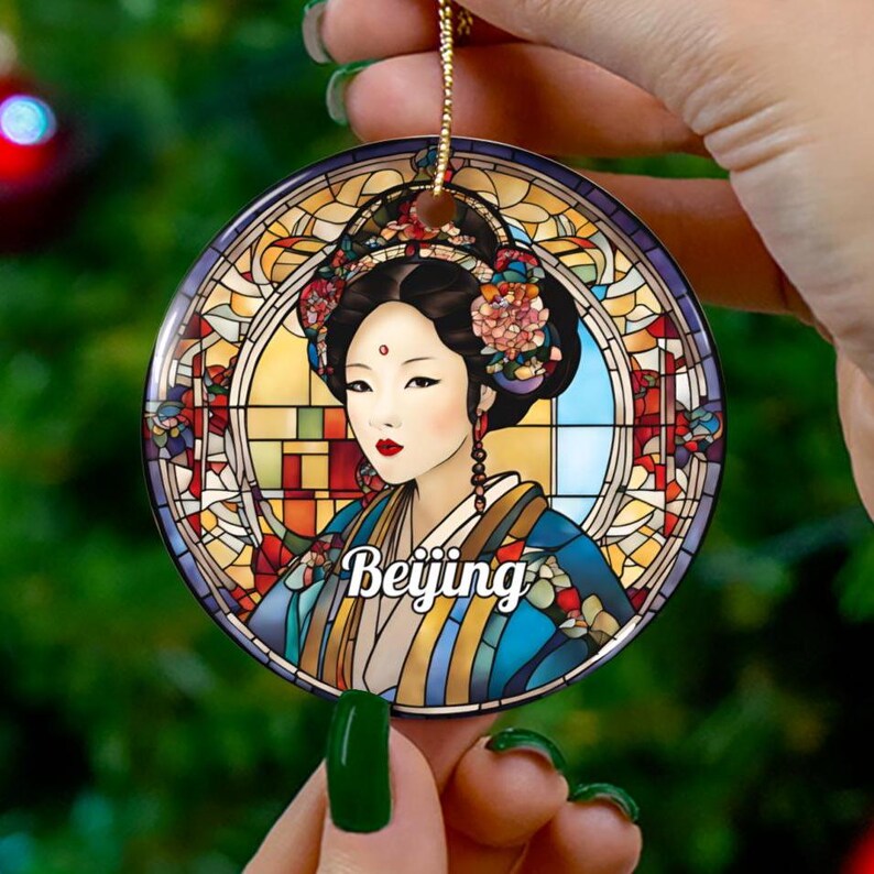 May include: A round, glass ornament with a stained glass design. The design features a woman with black hair and a blue and yellow robe. The woman is surrounded by a colorful stained glass frame. The text 'Beijing' is written in the center of the ornament.