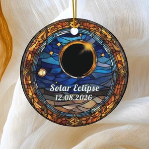 May include: A round ceramic ornament featuring a stained glass style design of a solar eclipse. The center shows a black circle surrounded by a glowing ring. The text reads Solar Eclipse and 12.08.2026. It hangs from a gold string.