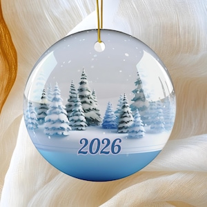 May include: A round, glossy, flat Christmas ornament featuring a winter landscape with snow-covered evergreen trees under a light blue sky. The year 2026 is printed in dark blue text at the bottom center. A gold string is attached to the top.