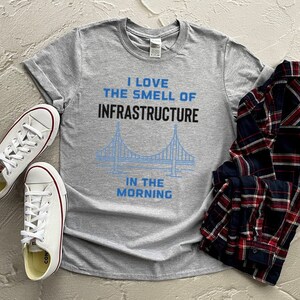 May include: A gray t-shirt with the text "I love the smell of infrastructure in the morning" and a blue outline of a bridge. The shirt is laid out on a gray surface with a plaid shirt and two white sneakers.