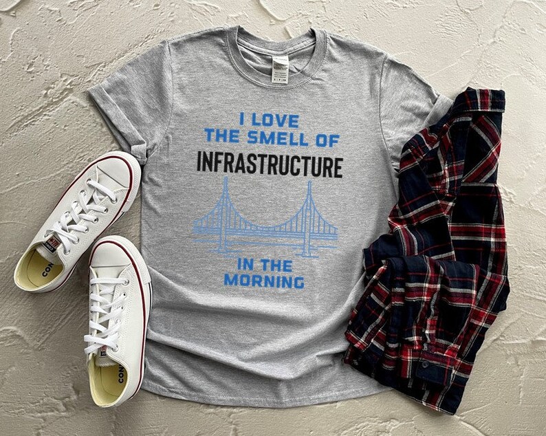 May include: A gray t-shirt with the text "I love the smell of infrastructure in the morning" and a blue outline of a bridge. The shirt is laid out on a gray surface with a plaid shirt and two white sneakers.
