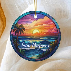 May include: Round ceramic ornament featuring a colorful sunset over the ocean with a palm tree. The text "Isla Mujeres" is written in a blue script. The ornament has a gold string for hanging.