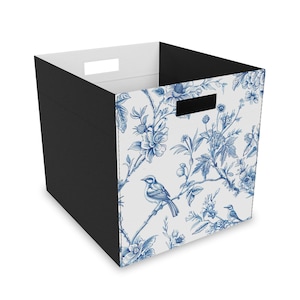 Puede incluir: Un cubo de almacenamiento cuadrado con exterior negro e interior blanco. Los lados presentan un patrón floral y de pájaros azul y blanco. Tiene dos asas rectangulares recortadas.