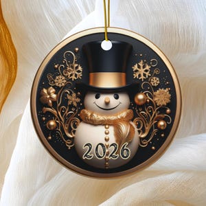 May include: A round black and gold Christmas ornament featuring a central snowman wearing a black top hat with a gold band. The design includes gold filigree, snowflakes, and the year 2026 printed in a decorative font. A gold cord is attached at the top.