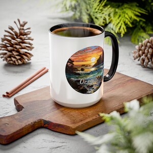 May include: White ceramic mug with a black handle and rim. The mug features a colorful oval design with a sunset over the Oregon coast and the word "Oregon". The mug is on a wooden cutting board.