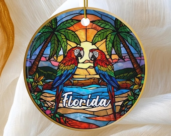 May include: Round ceramic ornament with a stained-glass design featuring two colorful parrots perched on a branch. The background includes palm trees, a sunset, and the word "Florida". The ornament has a gold-colored edge and a gold string for hanging.