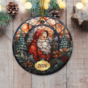 Puede incluir: Un adorno navideño redondo de cerámica con una ilustración estilo vidriera que muestra a Santa Claus con una larga barba blanca y abrigo rojo con capucha, rodeado de pinos y flores de pascua, con una etiqueta dorada que muestra el año 2026.