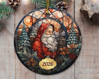 Op de afbeelding: Een ronde kerstornament van keramiek met een illustratie in glas-in-loodstijl van de Kerstman met een lange witte baard in een rode jas met capuchon, omringd door dennenbomen en kerststerren, met een gouden label met het jaartal 2026.