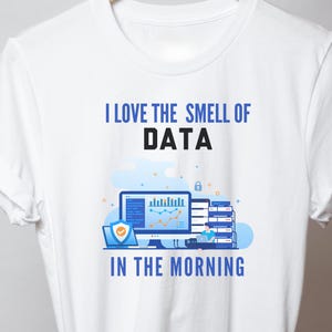 Pode incluir: Uma t-shirt branca de manga curta com uma impressão gráfica com o texto I LOVE THE SMELL OF DATA IN THE MORNING. O design inclui um monitor de computador com gráficos de dados, um portátil com um ícone de escudo e um servidor com uma pessoa.