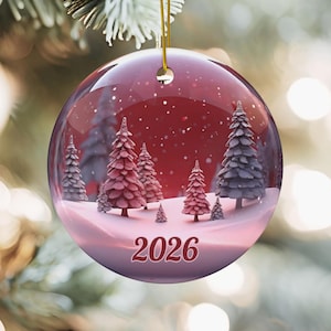 May include: A round, glossy ceramic Christmas ornament featuring a winter landscape with snow-covered pine trees against a deep red background. The design includes white falling snow and the year 2026 printed in a bold, dark red font at the bottom.