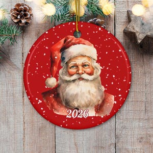 May include: A round red ceramic Christmas ornament featuring a detailed illustration of Santa Claus with a white beard and red hat. The background is a deep red with white falling snow accents. The year 2026 is printed in white at the bottom.