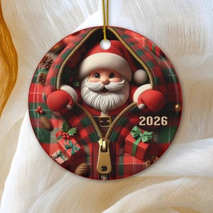 Puede incluir: Un adorno navideño redondo de cerámica con un gráfico de Santa Claus asomándose a través de una cremallera en una tela de cuadros rojos y verdes. El diseño incluye pequeños regalos, piñas y el año 2026 impreso en blanco a la derecha.