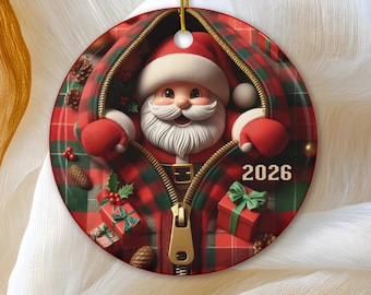 Op de afbeelding: Een ronde keramische kerstornament met een afbeelding van de Kerstman die door een rits in een rood-groene ruitjesstof kijkt. Het ontwerp bevat kleine cadeautjes, dennenappels en het jaartal 2026 in het wit aan de rechterkant.