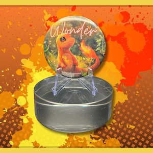 Peut inclure: Un bouton rond avec un dos en plastique transparent. Le bouton présente une illustration colorée d'une petite créature orange avec une queue et le mot "Wonder" écrit en cursive blanche.