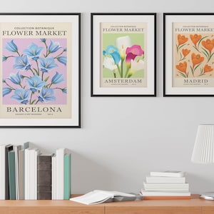 Puede incluir: Tres impresiones enmarcadas con un tema bot&aacute;nico. Cada impresi&oacute;n presenta una flor diferente y el nombre de una ciudad. La primera impresi&oacute;n muestra flores de campanilla azul y el texto "COLLECTION BOTANIQUE FLOWER MARKET BARCELONA". La segunda impresi&oacute;n muestra lirios calla blancos, rosas y azules y el texto "COLLECTION BOTANIQUE FLOWER MARKET AMSTERDAM". La tercera impresi&oacute;n muestra flores de azafr&aacute;n naranja y el texto "COLLECTION BOTANIQUE FLOWER MARKET MADRID".