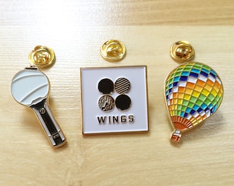 Puede incluir: Tres pines de oro con diferentes diseños. El primer pin es una luz de palo blanca y negra con la palabra "BTS" en el mango. El segundo pin es un cuadrado blanco con un logotipo negro y la palabra "WINGS". El tercer pin es un globo aerostático colorido.