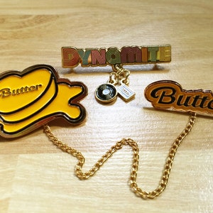 May include: Three gold-colored enamel pins with chains. The first pin says "Dynamite" in rainbow colors. The second pin is yellow and says "Butter" with a stylized butter pat design. The third pin is also yellow and says "Butter" in a curved font.