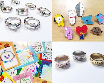 Puede incluir: Una colección de accesorios con temática BT21 que incluye anillos plateados con dijes de personajes, pines de esmalte coloridos con varias figuras, pegatinas holográficas con contornos de personajes y tres bandas metálicas anchas con iconos grabados.