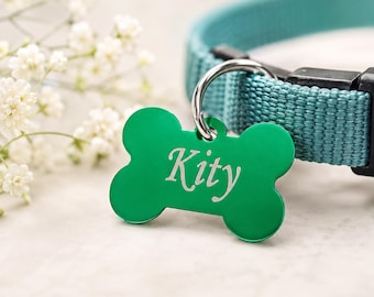 May include: A green, bone-shaped pet tag with the name "Kity" in white script. The tag hangs from a silver ring, attached to a teal collar. White flowers are in the background.