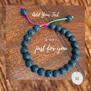 Könnte beinhalten: Schwarzes Lava-Stein-Armband mit einem bunten Schnurschluss und einem silbernen Anhänger mit dem Buchstaben "M". Das Armband trägt den Text "Ein Geschenk nur für dich" auf der Holzoberfläche, auf der es liegt.
