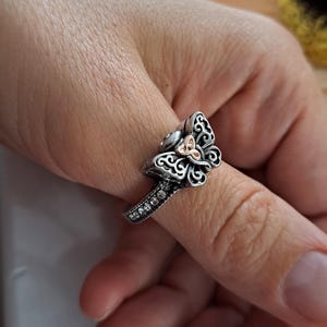 May include: Silver ring featuring a decorative floral design with a small gold-colored trinity knot in the center. The ring band is adorned with small, clear gemstones. The ring is worn on a finger.