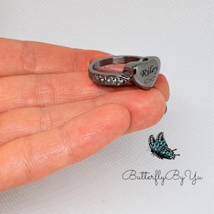 Könnte beinhalten: Ein schwarzer herzförmiger Ring mit einem silbernen Band und klaren Zirkonia-Steinen. Das Herz trägt die Gravur "Riley".