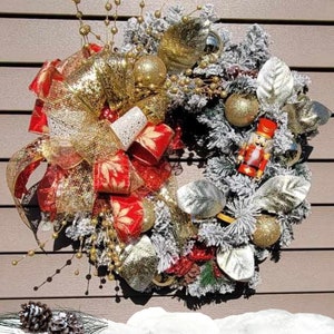 May include: A Christmas wreath with a red and gold bow, gold ornaments, and a nutcracker figurine. The wreath is decorated with white flocked pine branches and gold glitter.