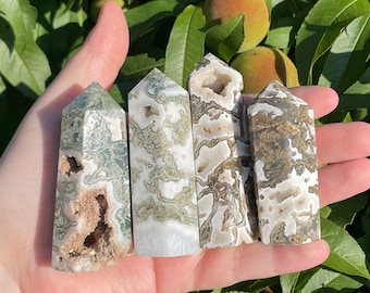 May include: Five green and white agate crystal points, also known as moss agate, are held in a hand. The points are various shades of green and white with a natural, rough texture.
