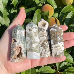 May include: Five green and white agate crystal points, also known as moss agate, are held in a hand. The points are various shades of green and white with a natural, rough texture.