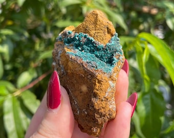 May include: A rough, brown, and green geode with a crystal formation inside. The geode is being held in a hand with red nail polish.