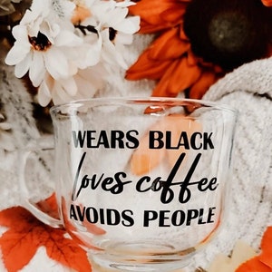 May include: A clear glass mug with a black inscription that reads "Wears Black Loves Coffee Avoids People". The mug is sitting on a white fuzzy surface with fall-colored flowers in the background.