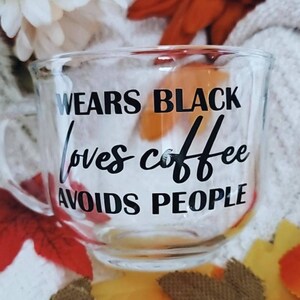 May include: A clear glass mug with a black inscription that reads "Wears Black Loves Coffee Avoids People".