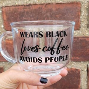 May include: A clear glass mug with a black inscription that reads "Wears Black Loves Coffee Avoids People".