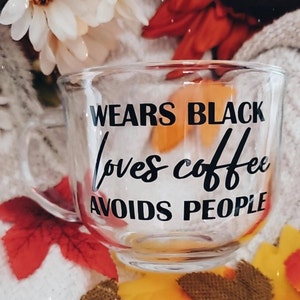 May include: A clear glass mug with black text that reads "Wears Black Loves Coffee Avoids People". The mug is sitting on a surface with fall-colored leaves.