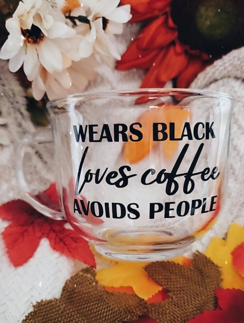 May include: A clear glass mug with black text that reads "Wears Black Loves Coffee Avoids People". The mug is sitting on a surface with fall-colored leaves.