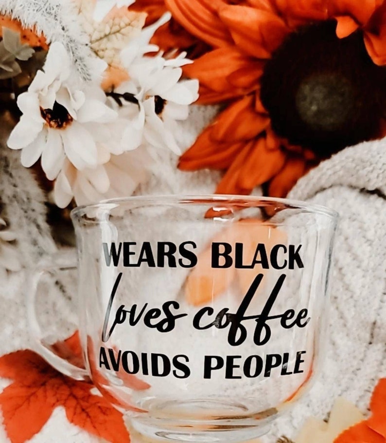May include: A clear glass mug with a black inscription that reads "Wears Black Loves Coffee Avoids People". The mug is sitting on a white fuzzy surface with fall-colored flowers in the background.