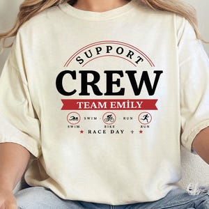 May include: A cream-colored short-sleeve t-shirt featuring a graphic design with the text SUPPORT CREW and TEAM EMILY in black and red. Below the text are three circular icons depicting a swimmer, a cyclist, and a runner with the labels SWIM, BIKE, RUN.